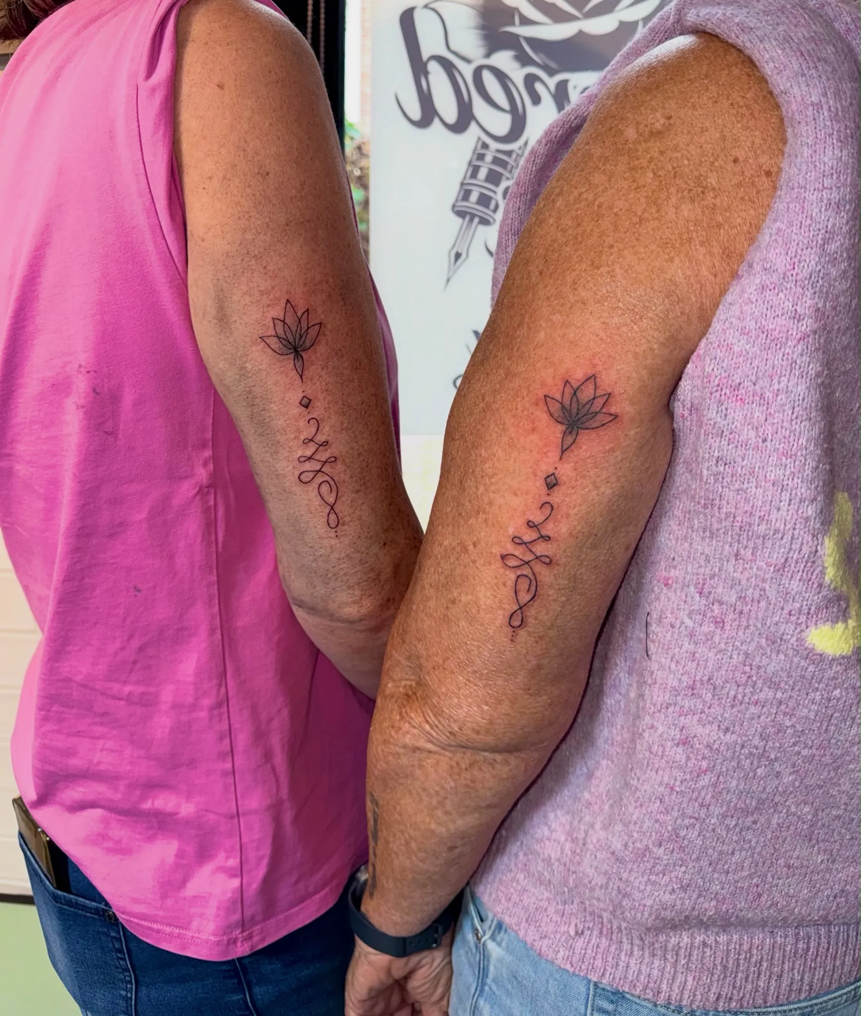 Twee bijpassende fineline tattoos van een lotus met unalome op de bovenarm van twee personen