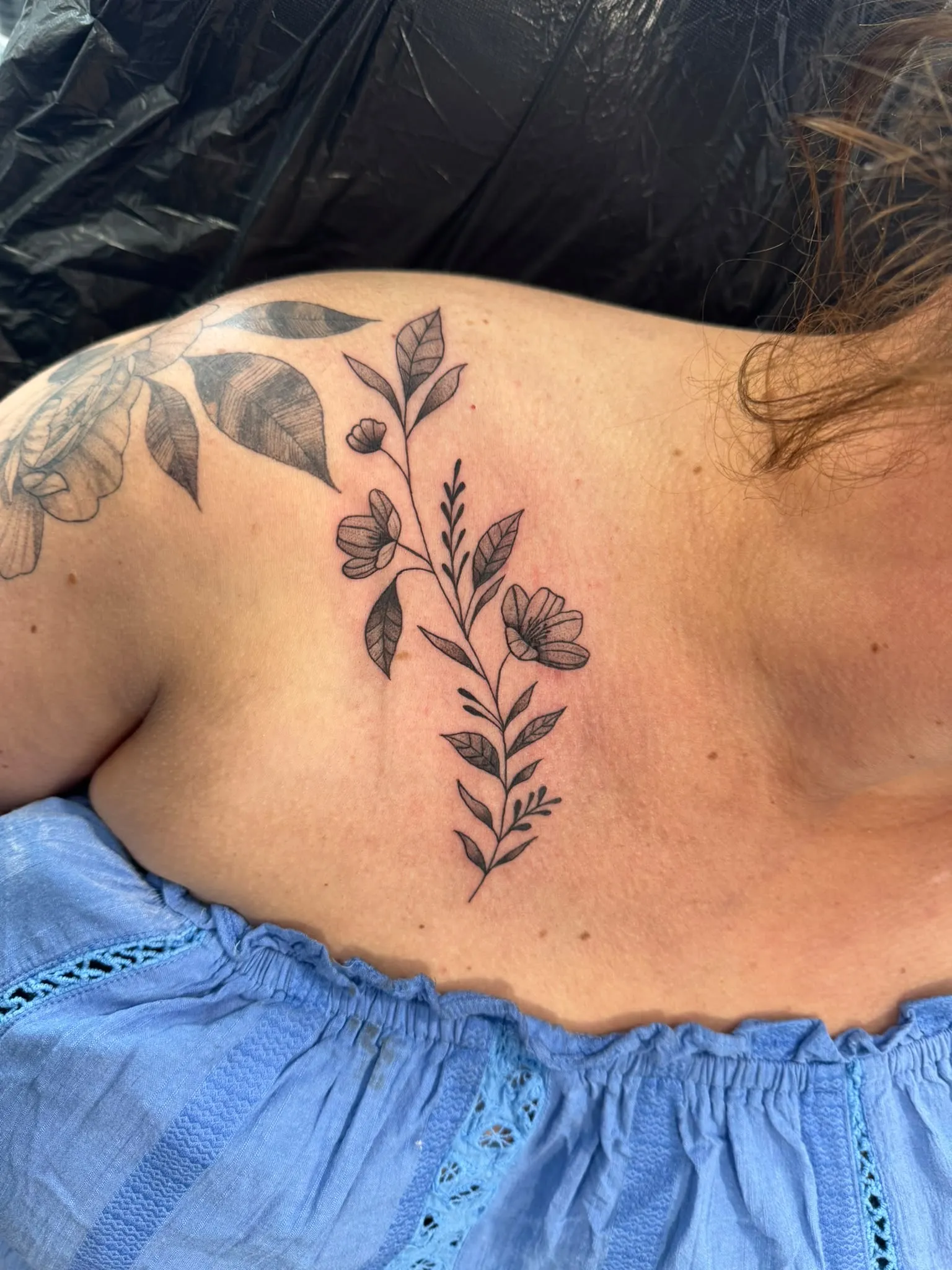Fineline floral tattoo van een bloemenrank met bladeren op het schouderblad, met zachte dotwork-schaduw