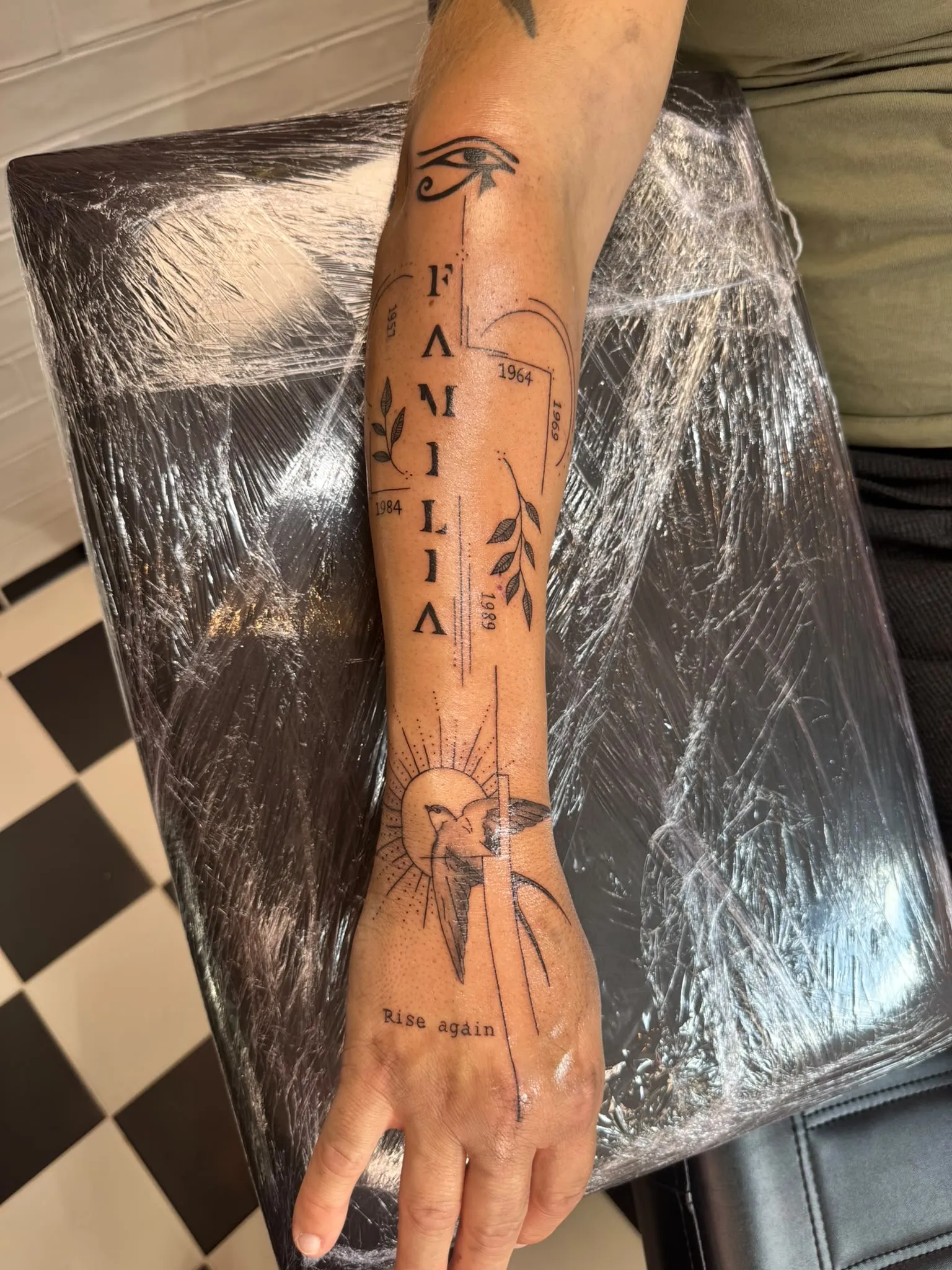 Tekst tattoo "Familia" met jaartallen, takjes, zwaluw en zon in fineline op de onderarm en hand