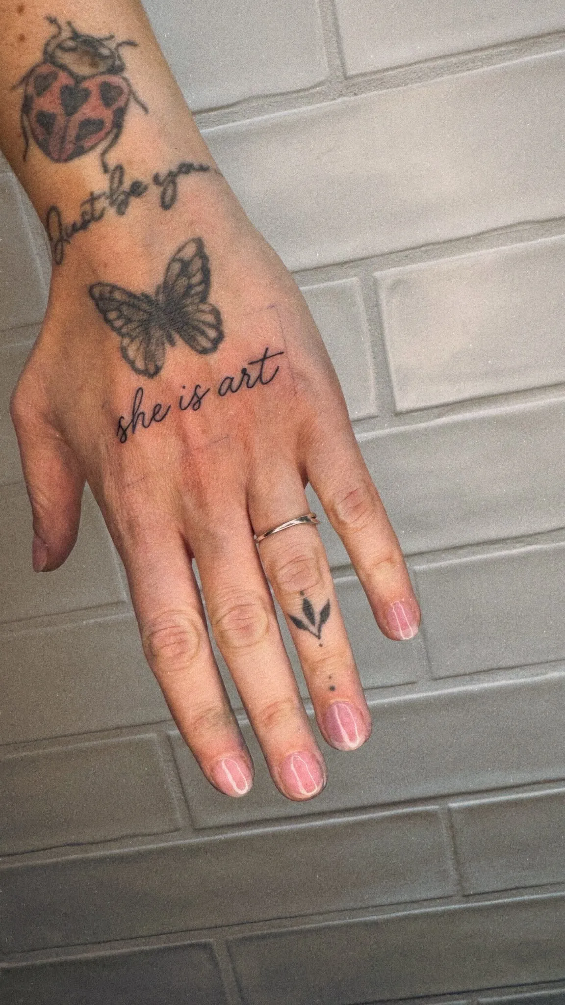 Kleine tekst tattoo "she is art" in handschrift op de hand, met een fineline blaadje op de vinger